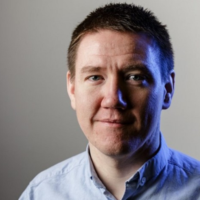 A man with short dark hair, dressed in a light blue button-up shirt, appears ready for training. The backdrop gracefully transitions from gray to blue.