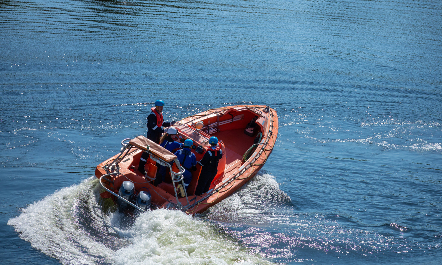 Four people wearing helmets and life jackets ride in an orange inflatable boat on the water.