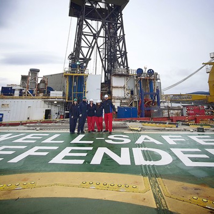 Four people in work uniforms stand on the deck of the offshore drilling rig "Well-Safe Defender", with industrial machinery in the background.