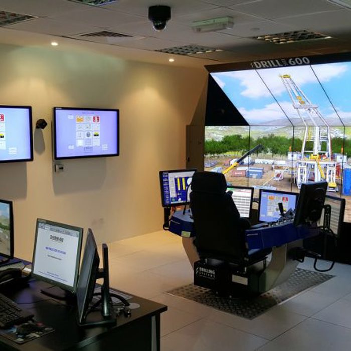 Control room featuring large screens and multiple workstations, including simulators for drilling operations, with on-screen visuals displaying various operational data and outdoor drilling imagery.