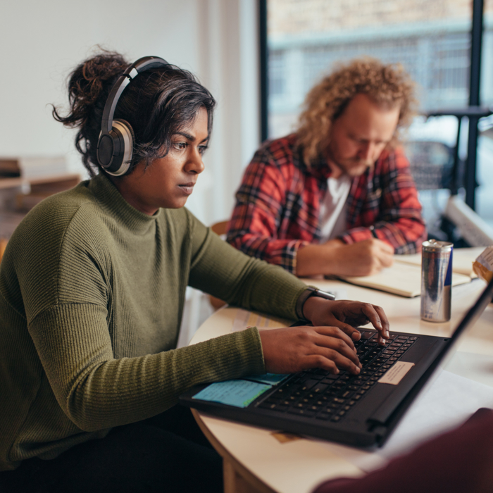 A person with headphones works on a laptop at a table, focused on training modules, while another person writes in the background.