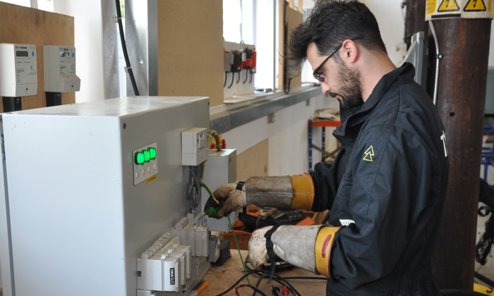 A man wearing protective gear works on an electrical panel, adjusting wires with precision in a utilities and construction training workshop.
