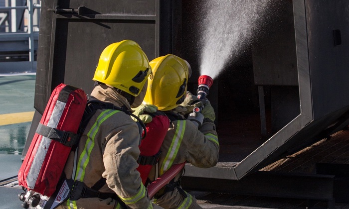 Firefighters in yellow helmets and protective gear spray water into a burning structure during a training exercise.