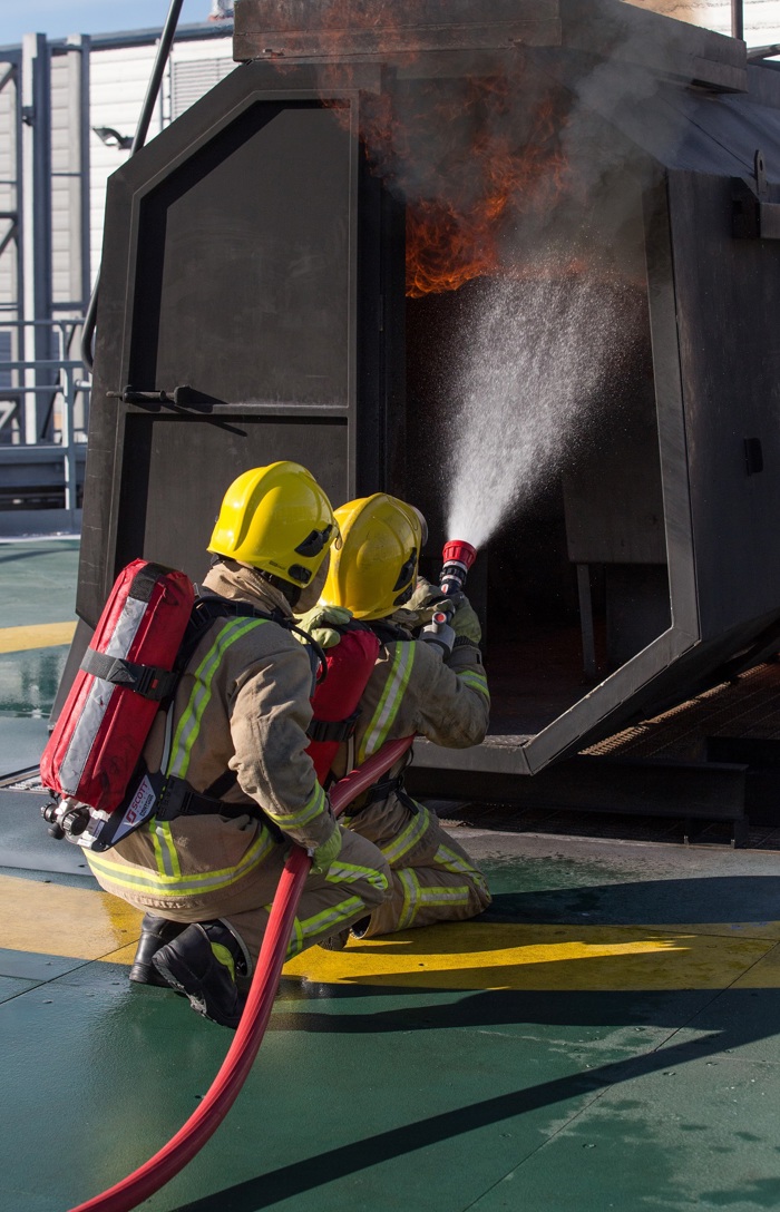 Firefighters in yellow helmets and protective gear spray water into a burning structure during a training exercise.