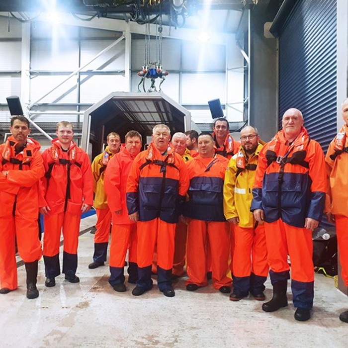 A group of people wearing orange and yellow safety attire stand indoors near a training pool facility.