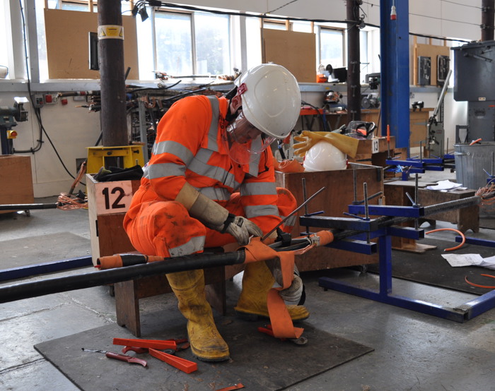A worker in orange protective gear and a white helmet repairs equipment in a workshop, surrounded by tools and supplies.