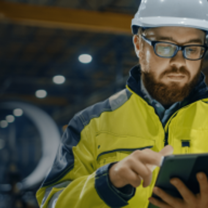 Man in high-visibility jacket and hard hat uses a tablet in an industrial setting.