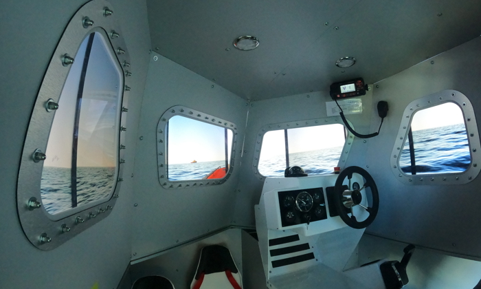 Interior view of a small, metal boat cabin with windows showing an ocean scene. The cabin contains navigation controls, a steering wheel, and seats.