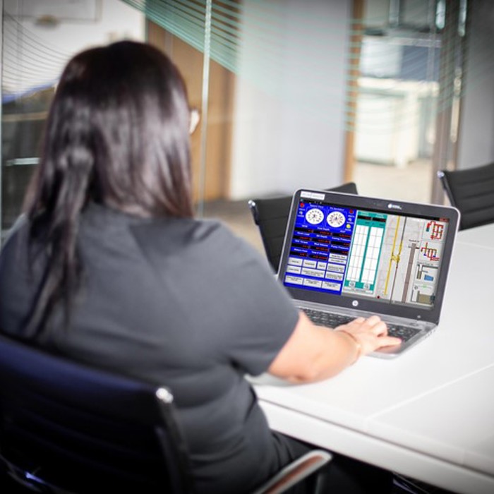 A person with long dark hair sits at a conference table, working on a laptop with a technical interface displayed on the screen.