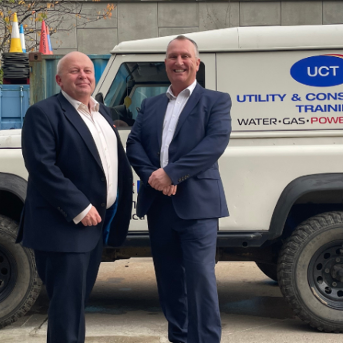 Two men in suits stand in front of a Utility & Construction Training vehicle, which has branding on the side indicating services related to water, gas, power, and highways.