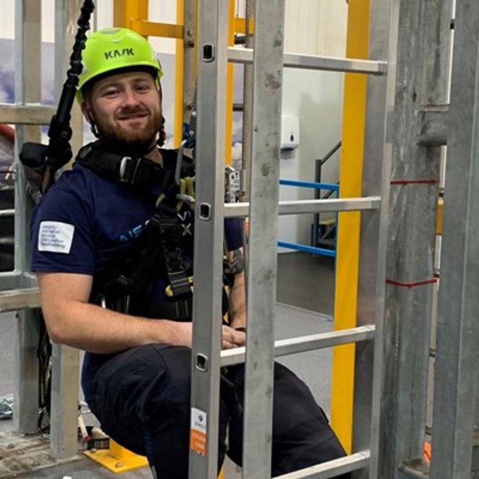 A man wearing safety gear and a helmet sits in a harness inside a metal structure indoors, with wind turbine models visible in the background.