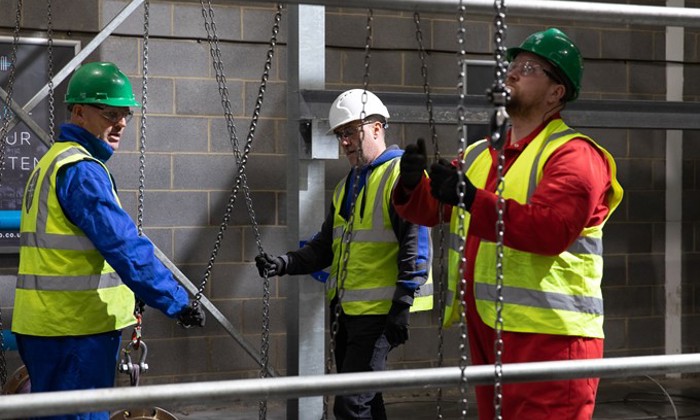 Three workers in high-visibility vests and hard hats operate chains and pulleys on a construction site.