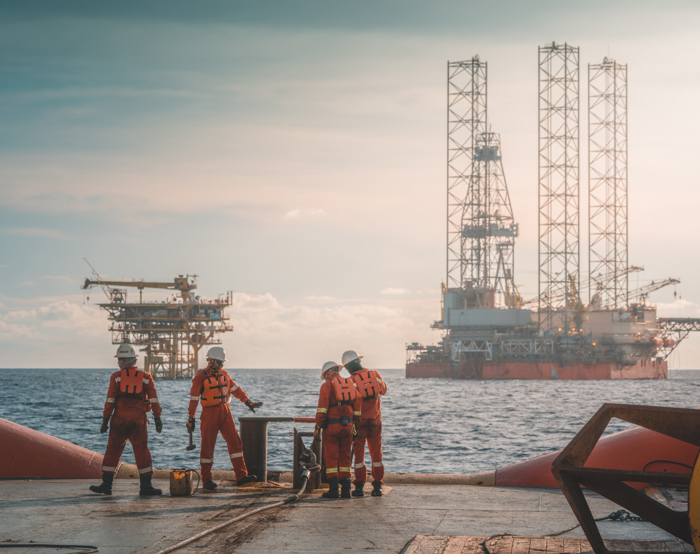 Four workers in safety gear stand on an offshore platform, honing their skills through hands-on training as they perform tasks, with two oil rigs visible in the background over the sea.