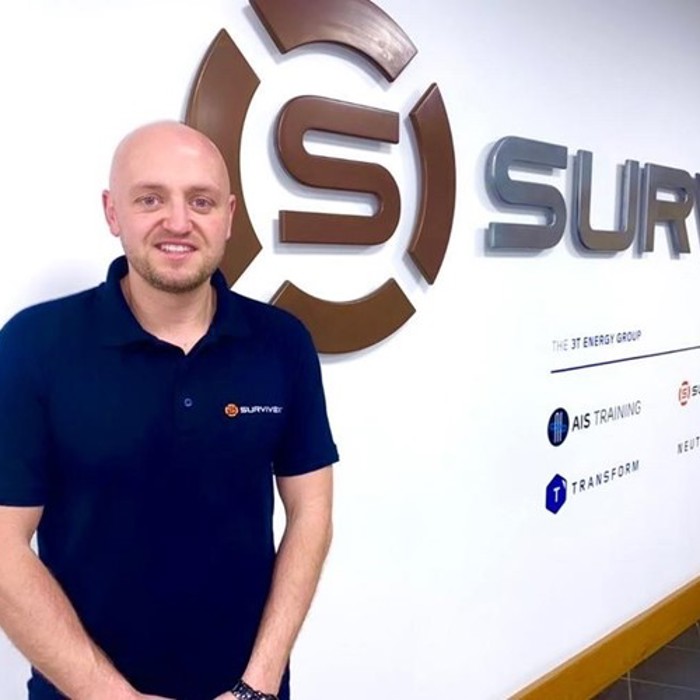 A person stands in front of a wall with the Survive logo and branding, dressed in a dark blue polo shirt with the same logo.
