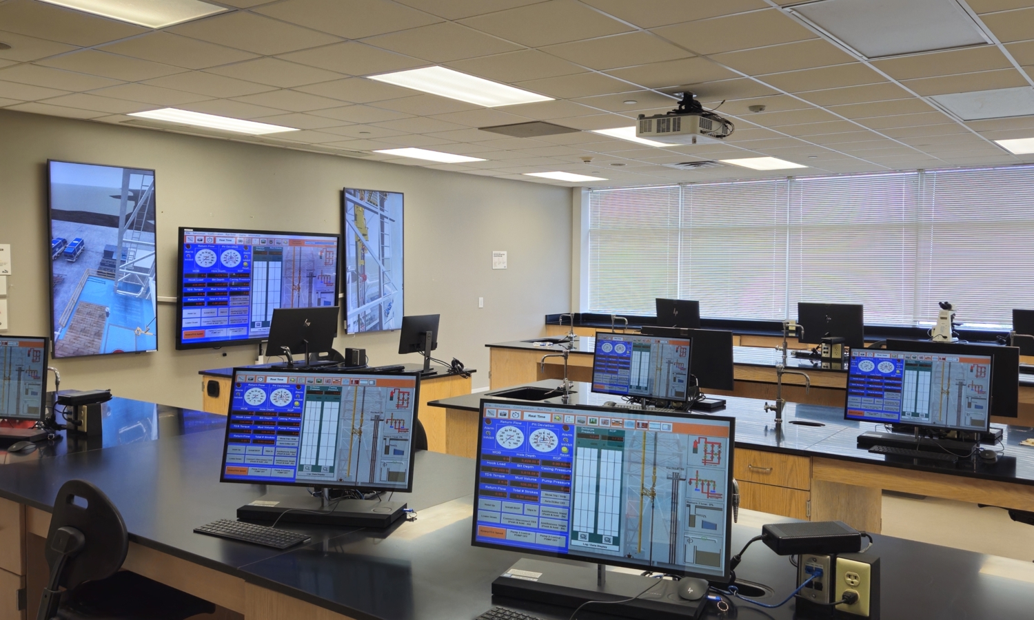 A modern control room with multiple workstations, each featuring monitors displaying control system interfaces. Large wall-mounted screens show surveillance camera feeds.