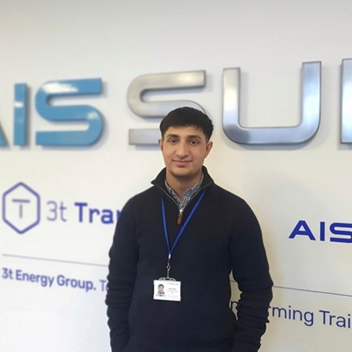 A person stands in front of a wall with the text "AIS Survivex" and "3t Energy Group" wearing a name badge and a navy blue sweater.