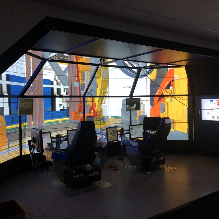 A modern simulation room with two control stations surrounded by large screen displays depicting an industrial environment.
