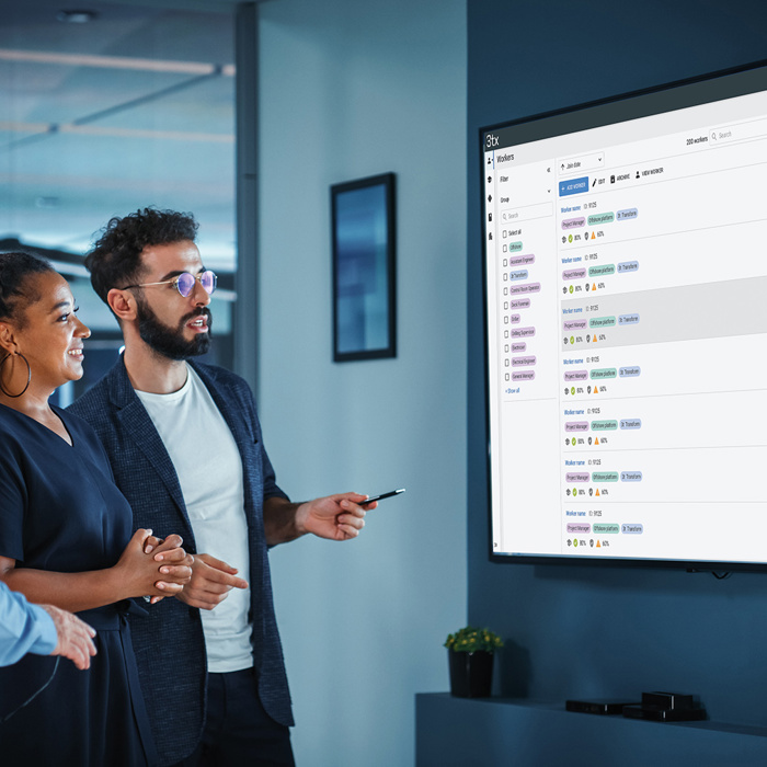Three people stand in an office, looking at a large screen displaying a training schedule. One person is pointing at the screen.
