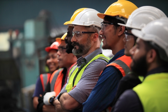 A group of workers wearing safety helmets and vests stands together, focusing intently on a training exercise just out of view.