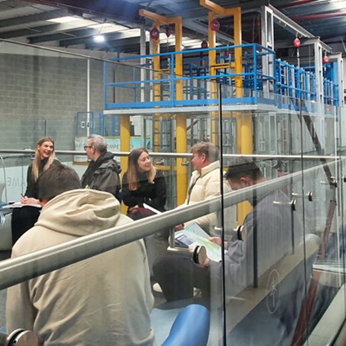 A group of people sits and converses in a modern industrial facility with glass partitions. Safety equipment and machinery are visible in the background.
