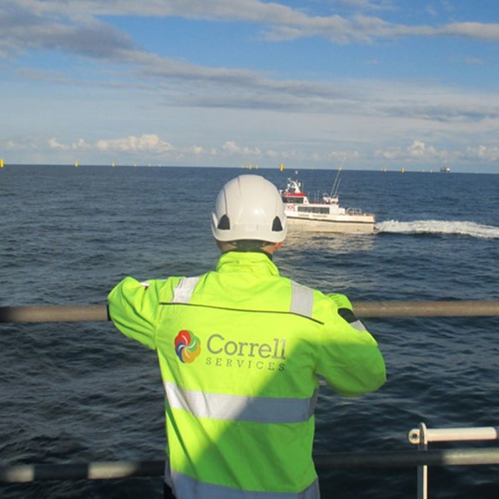 A person in a high-visibility jacket and helmet watches a boat in the ocean. The jacket has a "Correll Services" logo on the back.