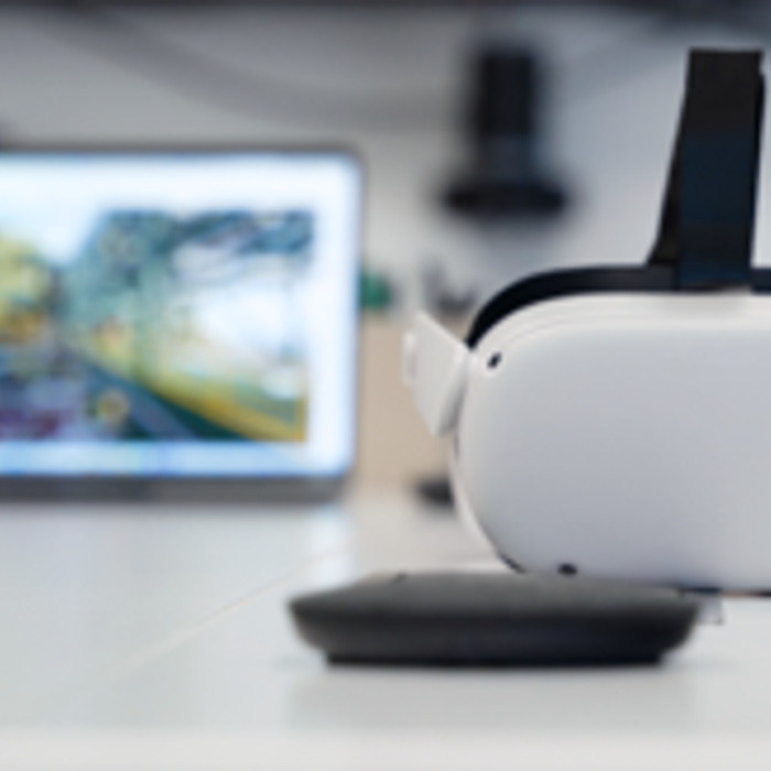 A VR headset and a controller are placed on the table in front of a blurred laptop displaying a landscape image, creating an immersive atmosphere that hints at virtual training possibilities.