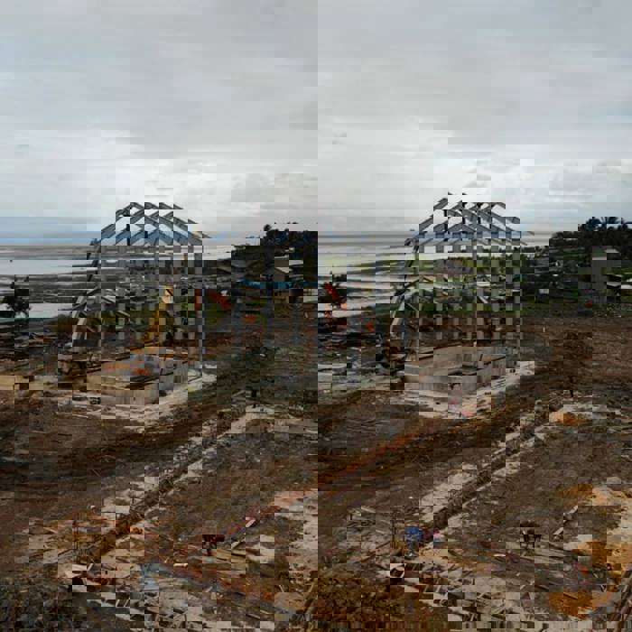 Aerial view of a construction site with steel framework in the center, surrounded by workers and machinery. The site is near a body of water and a small green building.