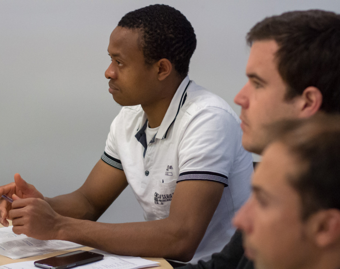 Three people are seated at a table, intently focusing on something out of view—perhaps a training session. One wears a white shirt while the others are partially visible. Papers and a phone lie scattered across the table.