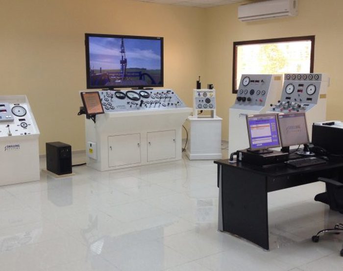 A control room with multiple consoles, a computer workstation, and large screens displaying data. Equipment includes dials, gauges, and control panels, all organized in a spacious, well-lit environment.