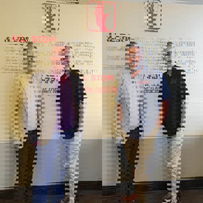 Two men stand confidently in front of a wall adorned with the text "ALL STOP! Survival & Safety Training." One dons a sharp blazer while the other opts for a sleek black shirt, embodying the essence of professional training preparation.