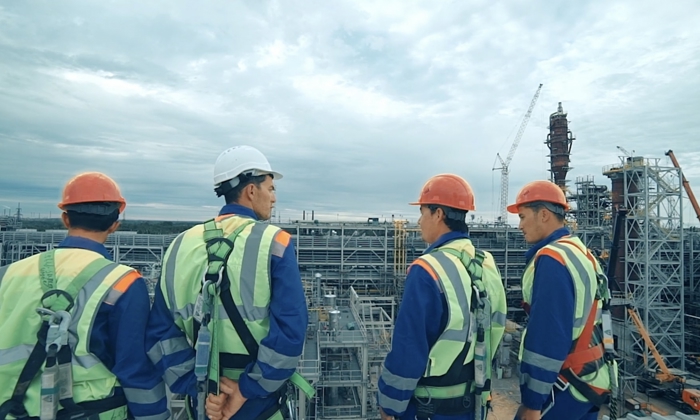 Four construction workers in safety gear discuss at an industrial worksite, with cranes and steel structures visible in the background under a cloudy sky.