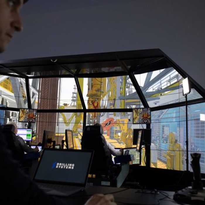 A person monitors multiple computer screens displaying drilling simulation software in a control room setting. The background shows a simulated drilling rig environment.
