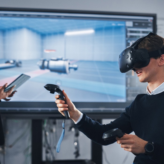 A person wearing a VR headset interacts with controllers while another person operates a tablet in front of a large screen displaying a 3D model in an office setting.