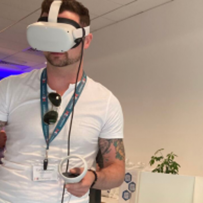A person wearing a white VR headset and holding controllers stands in a room with white walls and a television screen. They wear a white t-shirt and a lanyard, and have tattoos on their left arm.
