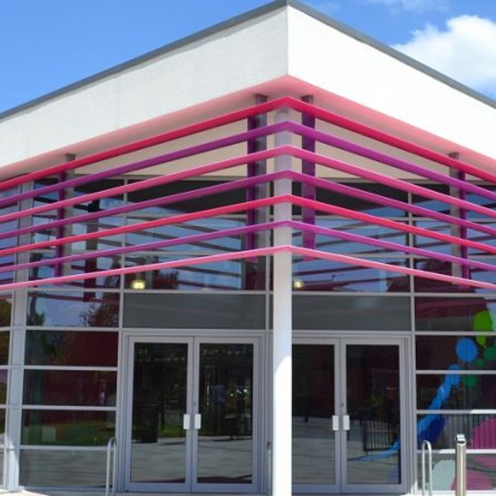 A modern building with pink accents and glass walls under a blue sky. "The Bourne Academy" is written on the upper edge of the building.
