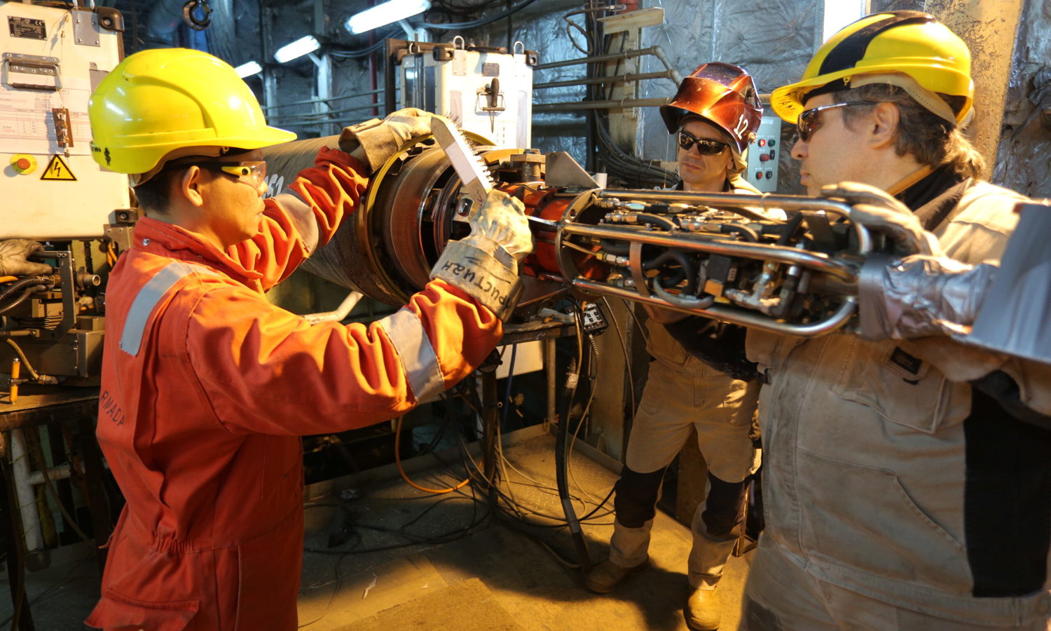 Three workers in protective gear are participating in training, collaborating in an industrial setting while handling machinery parts with tools and equipment around.