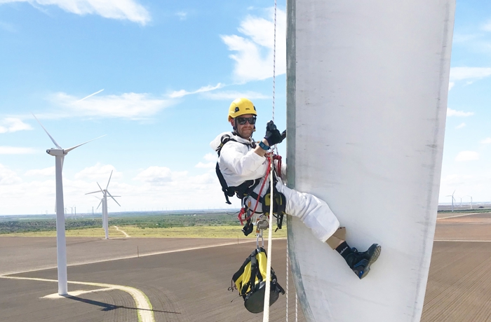 A worker in safety gear, including a helmet and harness, is suspended on a rope while performing maintenance on a large wind turbine blade with several wind turbines in the background, highlighting the military to wind transition in skilled labor.