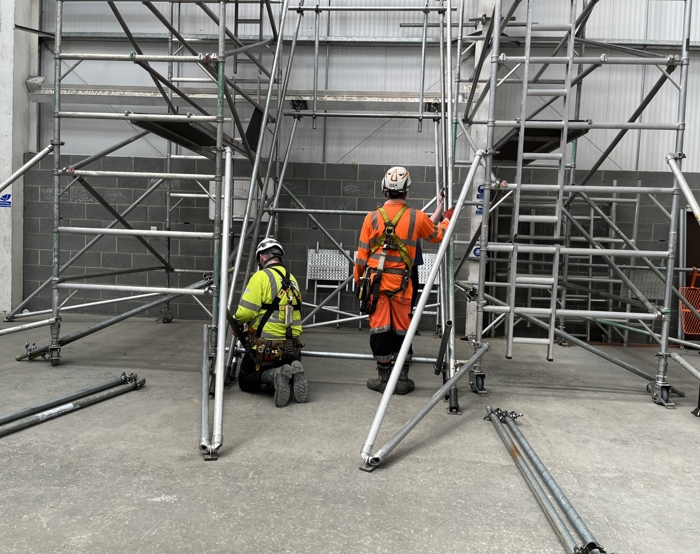 Two workers in safety gear carefully assemble scaffolding in an industrial setting, demonstrating precise training and expertise.