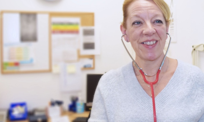 A medical professional with a stethoscope around her neck stands in a well-lit office with paperwork and a computer on a desk in the background.