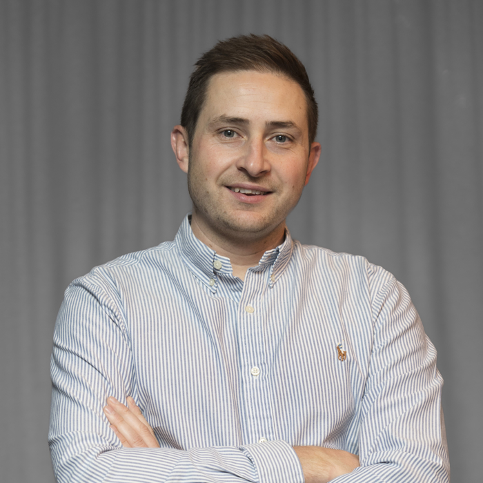 A man with short brown hair wearing a striped button-down shirt stands with arms crossed against a plain backdrop.