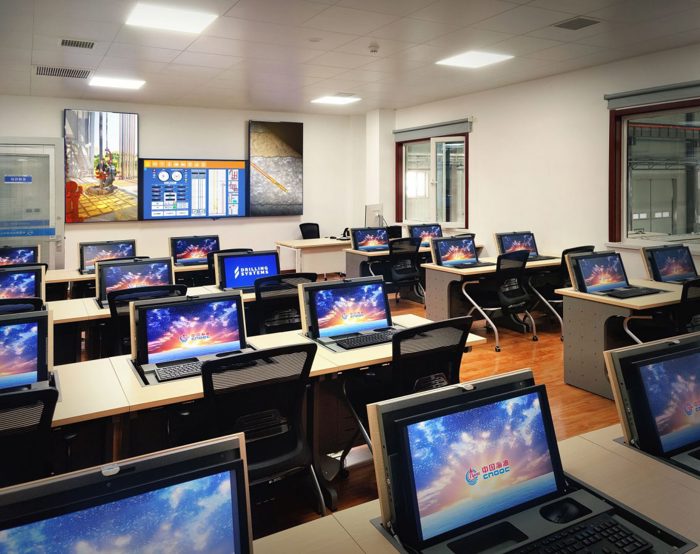 A modern classroom equipped with numerous workstations, each featuring a computer displaying a colorful screensaver. A large digital screen and whiteboard are visible at the front of the room.