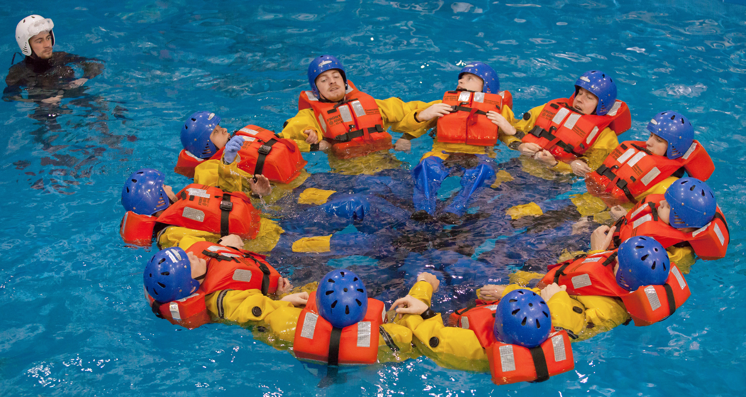 A group of people in life jackets and helmets form a circle in a pool as part of a training session. A person in black observes from the side.