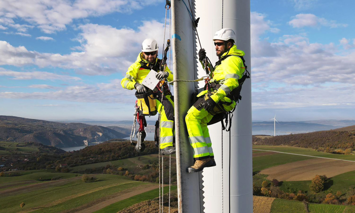Two workers in safety gear, demonstrating their training, inspect a wind turbine while harnessed on ropes, with a scenic landscape of hills and more turbines in the background.