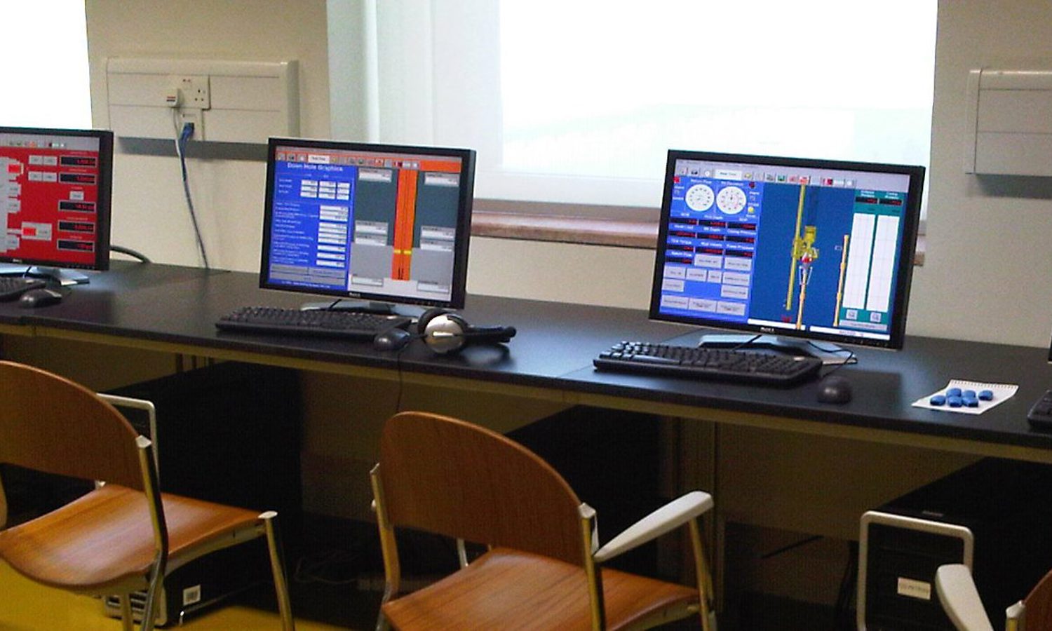A row of five computer workstations with various software interfaces displayed on the monitors. There are keyboards, mice, and headsets at each station, with wooden chairs facing the monitors.