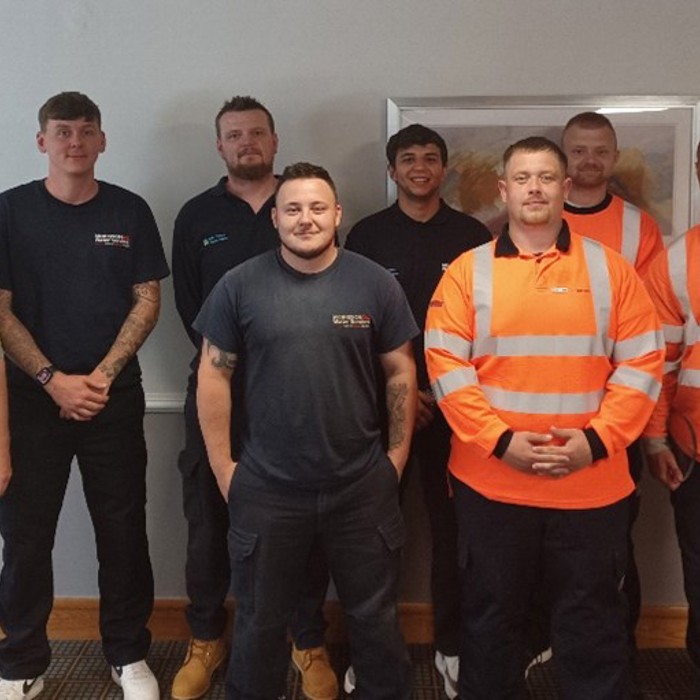 Eight men stand indoors, some in work uniforms, possibly gathering for a training session.