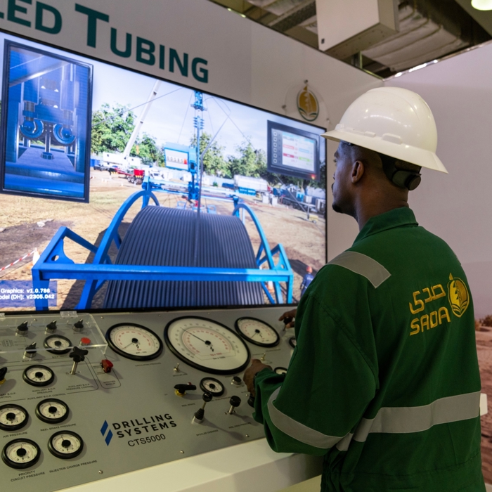 A person in a hard hat operates a sophisticated drilling simulation console with gauges, engaged in an advanced training session, as a large screen vividly displays an oilfield scene.