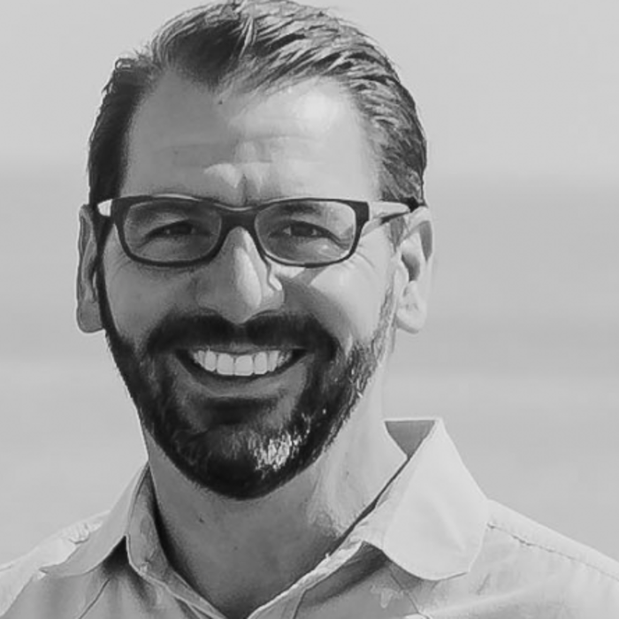 A man with short dark hair, a beard, glasses, and a light-colored shirt is smiling. The background shows a blurred expanse of water.
