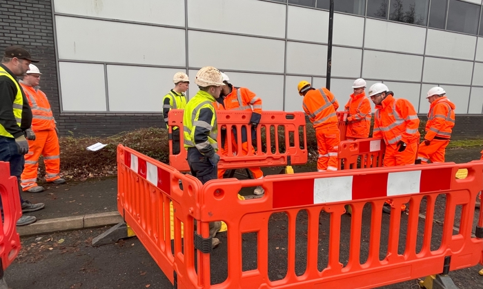 Construction workers in safety gear, undergoing training, stand near orange barriers on a road beside a building.