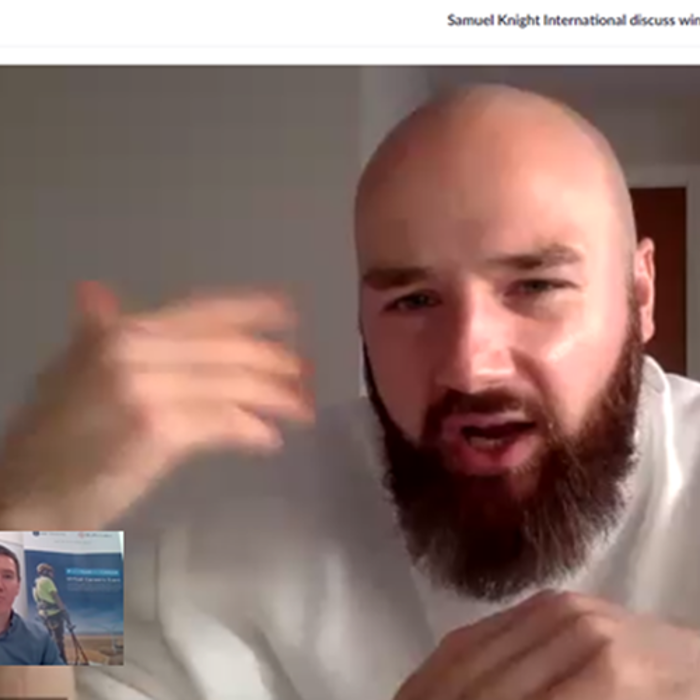 Two men are shown in a Zoom video call discussing a wind project, one speaking with an animated gesture, the other visible in a smaller window.