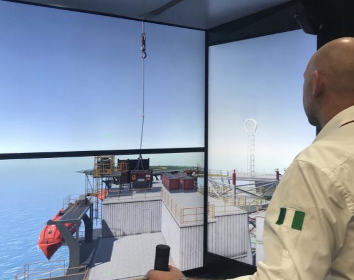 A person operates a crane simulator, controlling a hoist above an offshore platform displayed on large screens.